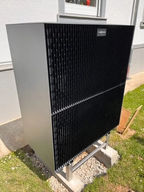 Dunkles Außengerät einer Wärmepumpe im Garten neben einer Hauswand